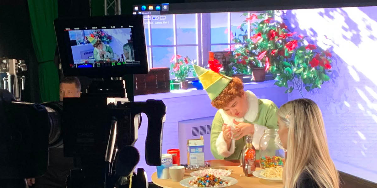 Actor in elf outfit sitting at a table with sweets and film crew and cameras filming