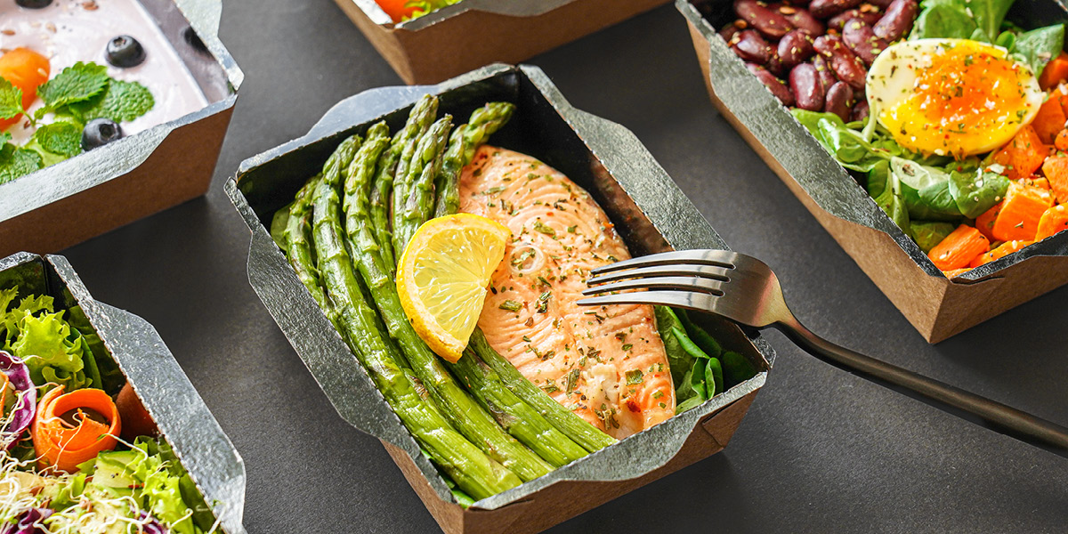 Boxed salmon and asparagus with fork surrounded by other healthy food boxed in takeaway boxes 