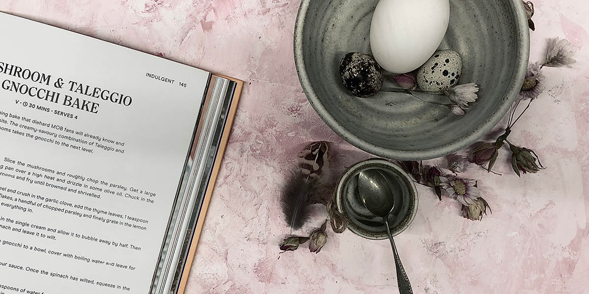 White and speckled egg in a brown bowl set against a pink background and set against a recipe book