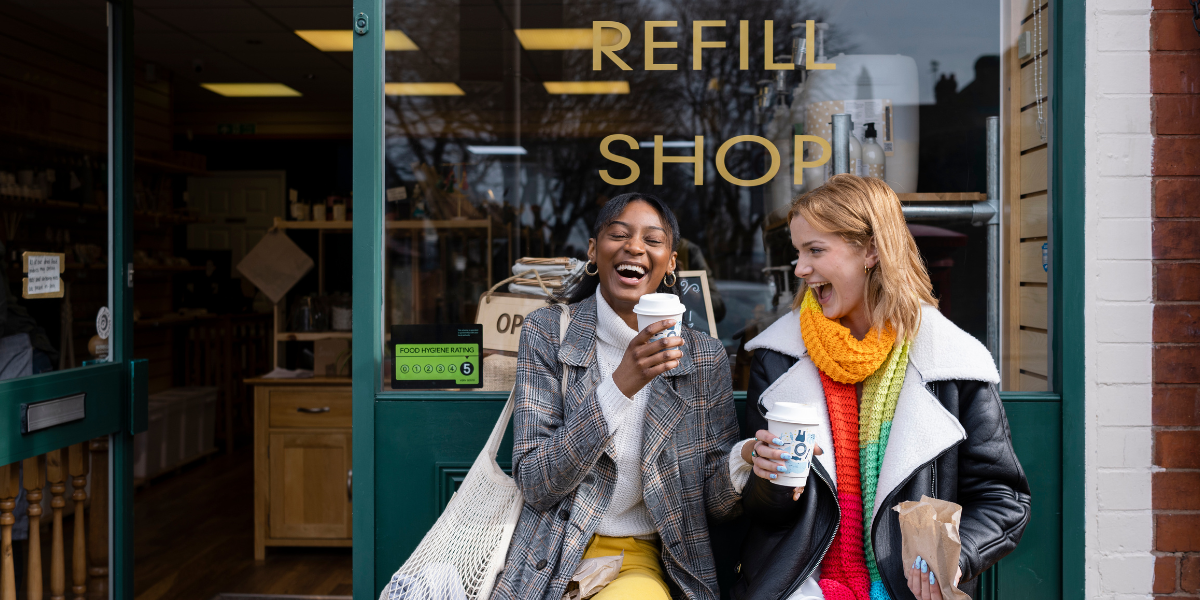 two women sitting outside a refill shop with coffee in their hands